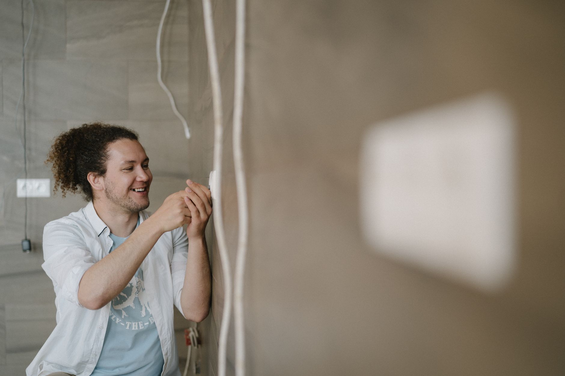 Un électricien avec des cheveux bouclés ajuste une prise à l'intérieur, souriant pendant qu'il travaille.